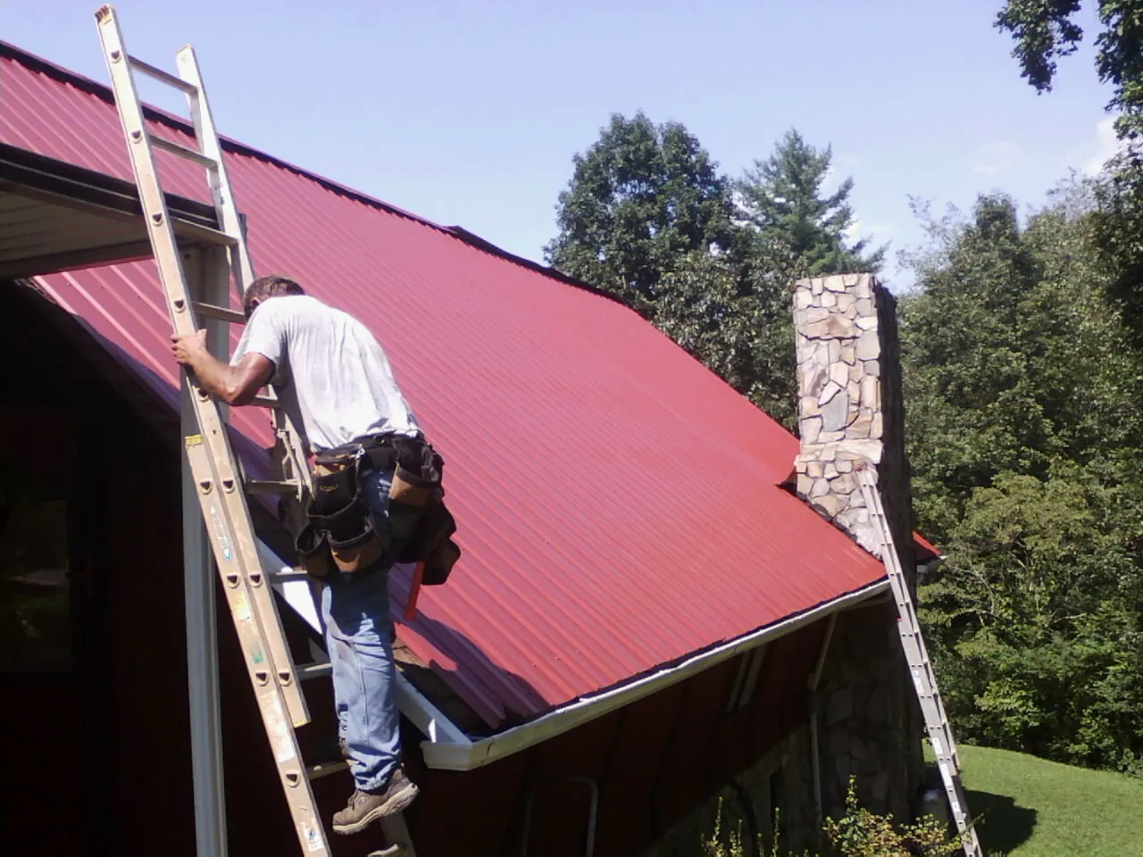 Roofer on a ladder beside a red metal roof and stone chimney — Soffit & Fascia Repair by Roofing Brothers in Spindale
